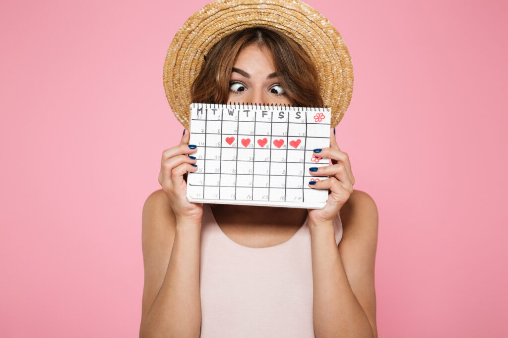 Portrait of a crazy funny girl in summer hat holding and hiding behind a periods calendar isolated over pink background