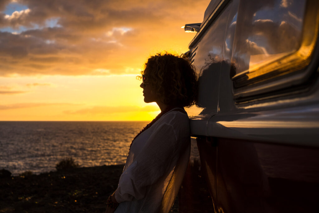 Travel and wanderlust lifestyle destination concept with beautiful woman in silhouette and red vintage van enjoying the coloured orange sunset in front of the ocean view - independence and people