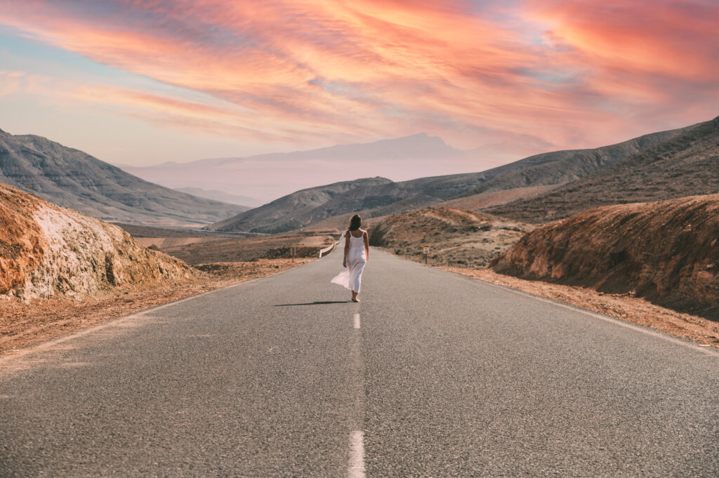 Back view full length of faceless female in white dress strolling on asphalt roadway in Canary Island in Spain between mountains under cloudy blue sky in countryside in daylight