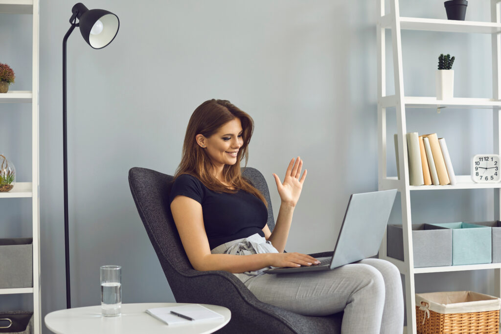 Woman waving hand has video call laptop computer online sitting in armchair at home. Remote work or training in the living room.